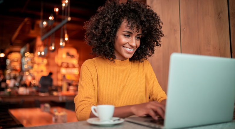 A foto mostra uma mulher sorridente usando um notebook ao lado de uma xicara de café, parece estar em uma cafeteria.