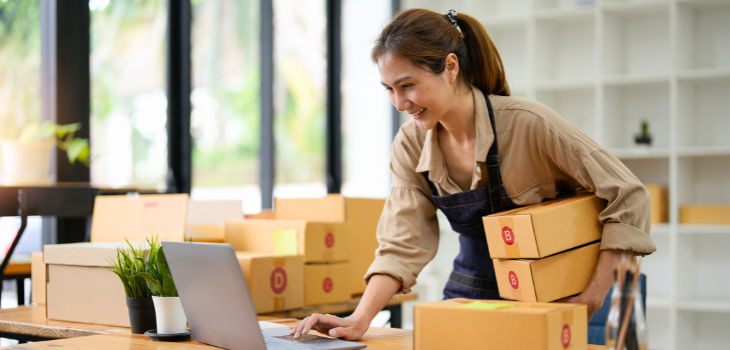 A imagem mostra uma mulher sorridente segurando algumas caixa para envio de produtos, ao mesmo tempo ela também está mexendo no computador.