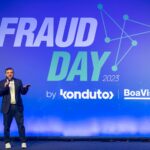 Foto do Lucas Guedes (homem vestindo terno azul, blusa branca e tenis branco) apresentando no palco do Fraud Day. No fundo azul está escrito "Fraud Day".
