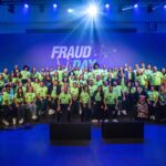 Foto colorida da equipe de organização do evento. A imagem mostra várias pessoas sentadas e de pé usando a camiseta do evento. No fundo está escrito "Fraud Day".